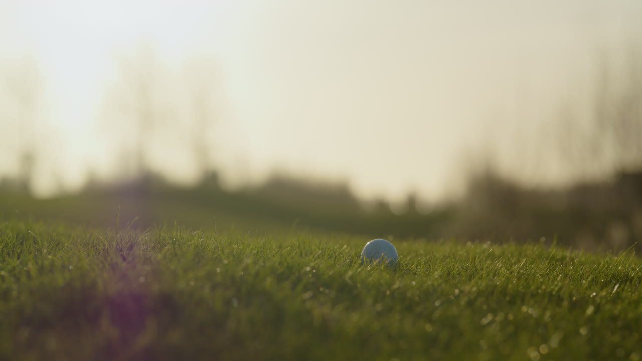 Close up of golf club hitting ball on green grass during golden hour. Slowmo, low POV