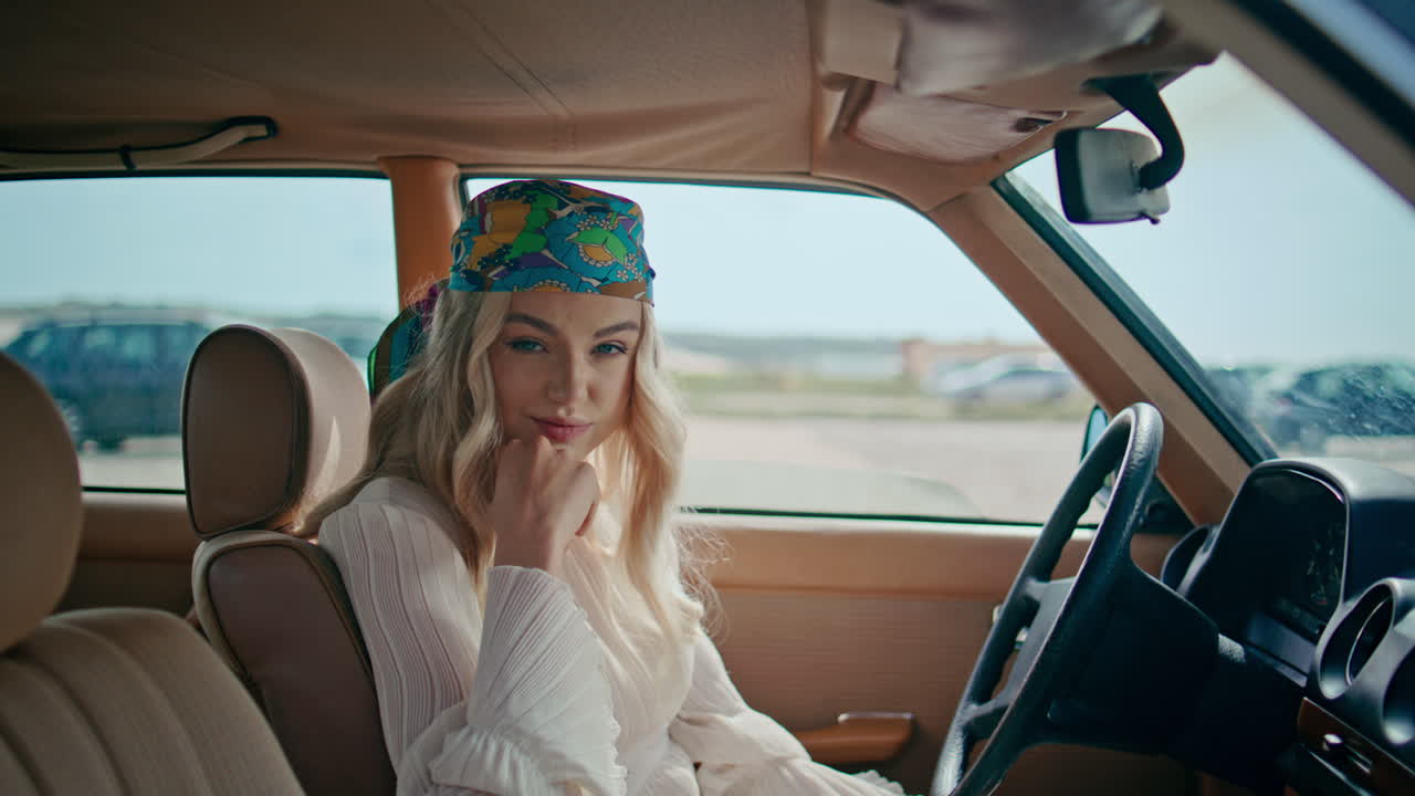 화려한 반다나를 입은 편안한 소녀가 자동차에 앉아 있습니다. 금발이 레트로 차로 포즈를 취하고 있습니다.