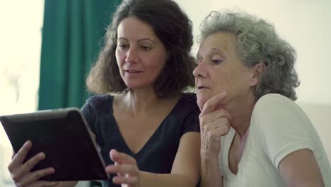 Two-smiling-women-sitting-at-table-and-using-tablet.