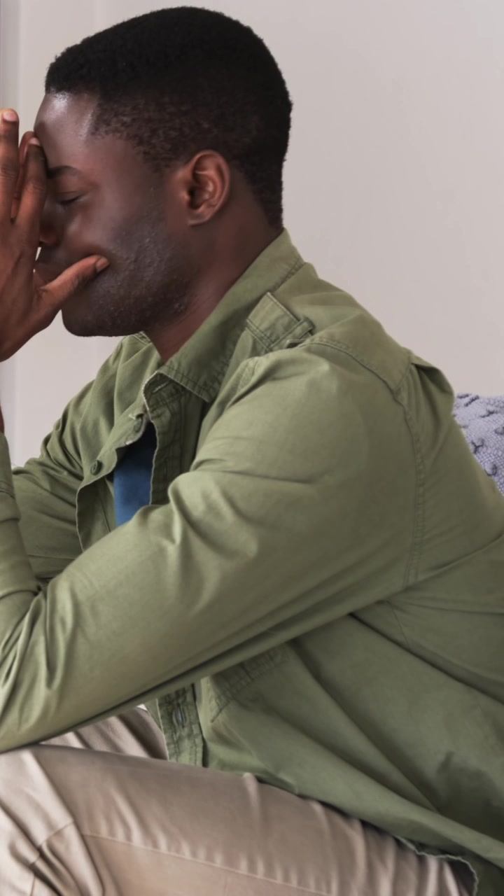Video verticale: Uomo afroamericano che si preme il viso e si stringe la testa cercando calma sul divano con una maglietta verde.