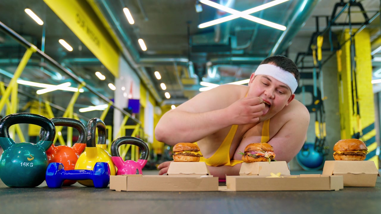 Uomo in sovrappeso che mangia fast food in palestra con attrezzature fitness