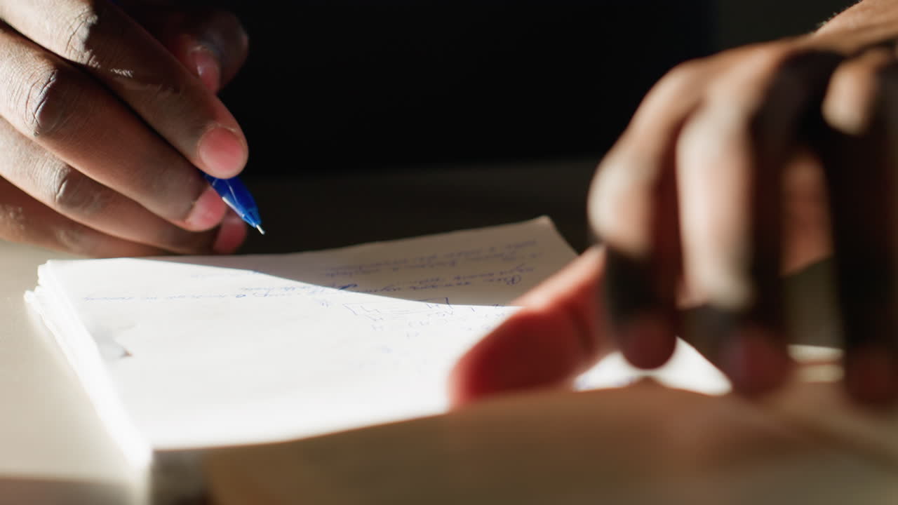 Close up of person writing notes in book with blue pen, focusing on hand, fingers, and paper under warm light, capturing act of recording thoughts or studying in quiet indoor environment