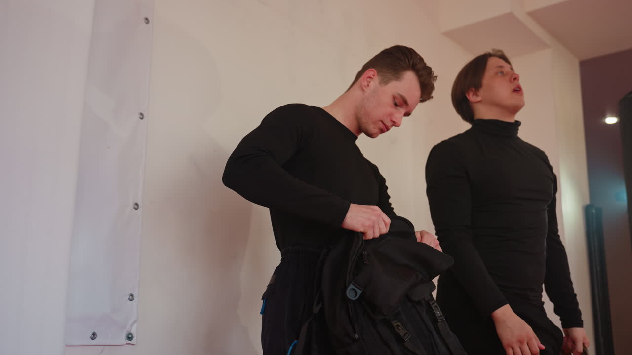Two men in black uniform standing up, one checking bag with intent focus, other looking upward with serious expression, scene suggesting discipline, awareness in training atmosphere