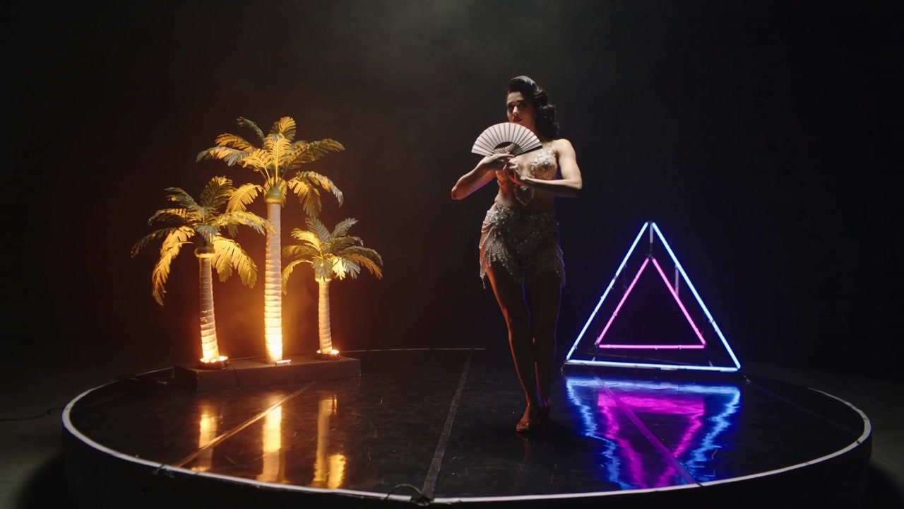 Ballerina che si muove incrociando le gambe, apre un ventaglio sul palco, indossa un abito scintillante, con triangolo al neon e palme color ambra