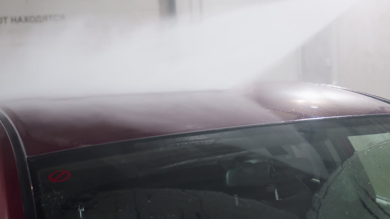 close up of red car roof being rinsed by high pressure wash spray with rising vapor against white wall with blurred inscription delivering dynamic wash action and glossy sheen on metal surface
