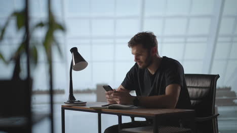 Lächelnder-Junger-Mann,-Der-Mobile-Apps-Nutzt,-Sitzt-Am-Tisch.-Männlicher-Tausendjähriger-Berufstätiger,-Der-Moderne-Smartphone-SMS-Nachrichten-Im-Büro-Hält.-Junger-Geschäftsmann-Nutzt-Hilfreiche-Mobile-Apps-Für-Seine-Geschäftszeit