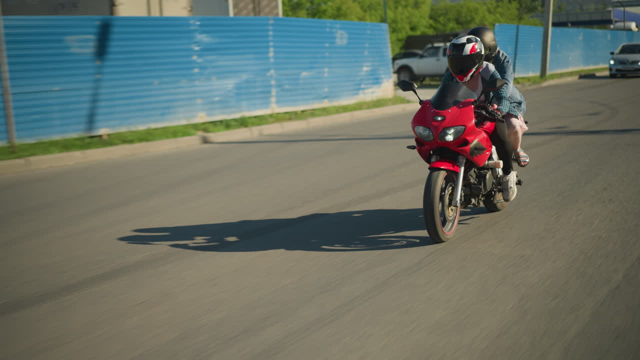 Two sisters on a red power bike ride along an urban road, passing a building and parked car seen through a zinc gate, an ash-colored car follows from behind, its headlamp on