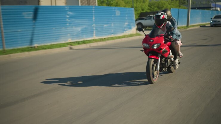 Two sisters on a red power bike ride along an urban road, passing a building and parked car seen through a zinc gate, an ash-colored car follows from behind, its headlamp on