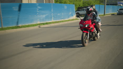 two sisters on a red power bike ride along an urban road, passing a building and parked car seen through a zinc gate, an ash-colored car follows from behind, its headlamp on