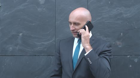 confident businessman standing at office building wall and talking on mobile phone