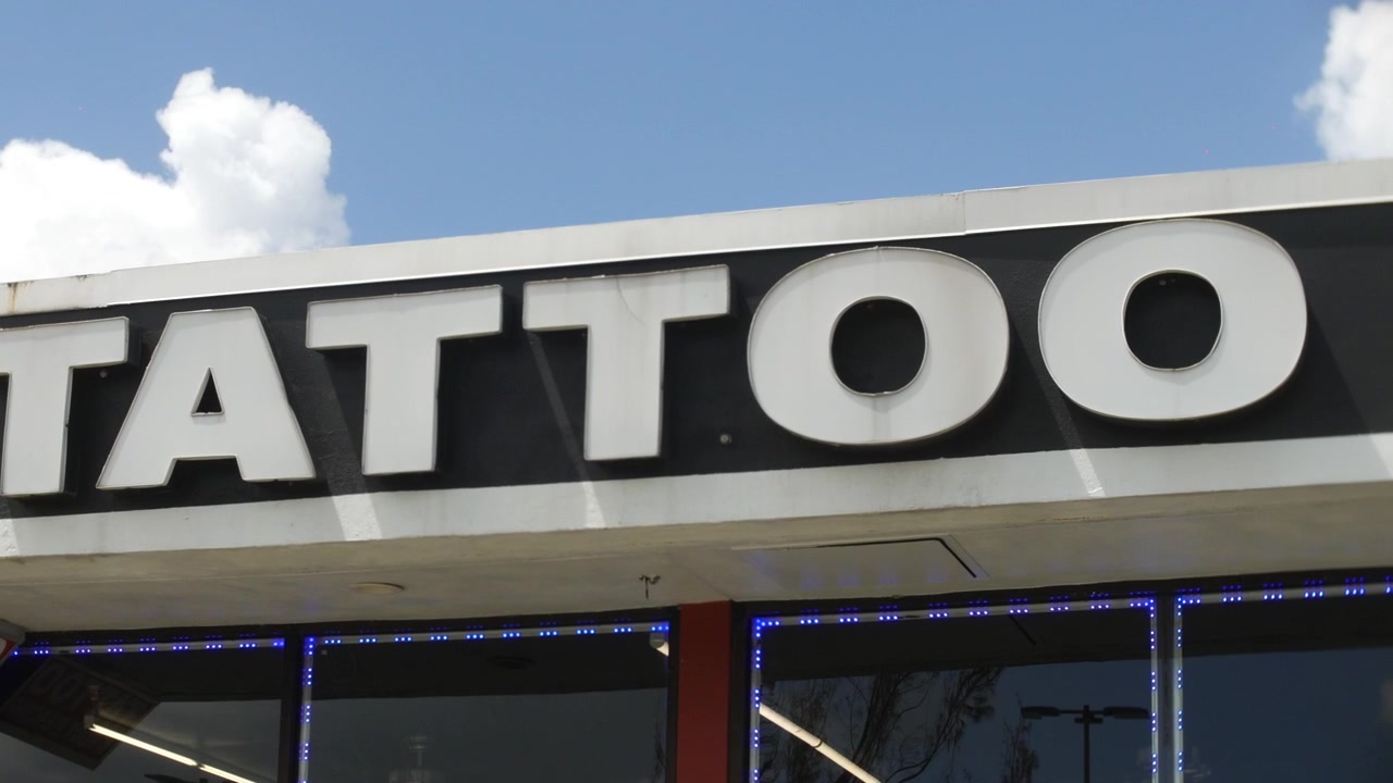 Insegna con la scritta TATTOO che occupa la facciata di un negozio sotto un cielo blu e nuvole. La vista ravvicinata mostra grandi lettere bianche, finiture nere, finestre di vetro e luci LED blu.