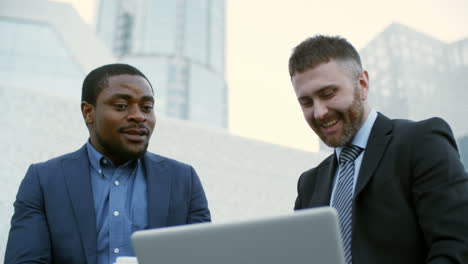 and businessmen sitting outdoors, looking at laptop screen and discussing some issues