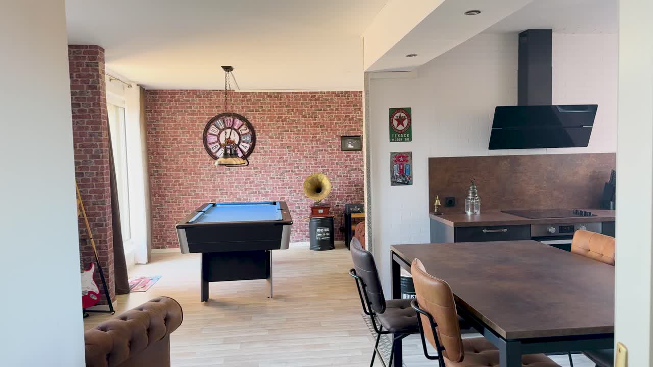 Camera pans through a modern apartment, revealing a billiard table, exposed brick wall, kitchen, and eclectic decor in bright natural light