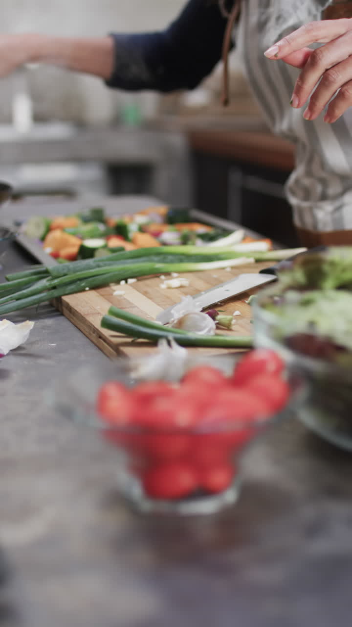 Vertical video of midsection of biracial woman in apron preparing meal in kitchen, slow motion