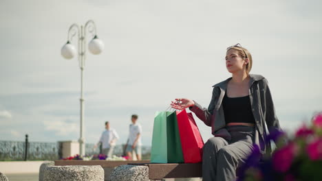 lady sits on an outdoor bench, placing her green and red shopping bags next to her, she adjusts the bags while carrying a black handbag, two people walk by in the background