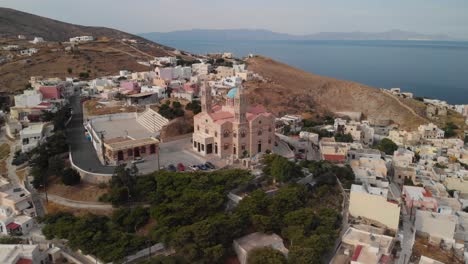 Vuelo-Aéreo-Hacia-Atrás-Desde-La-Hermosa-Iglesia-En-La-Cima-De-La-Colina-En-Syros,-Grecia-Con-El-Océano-Y-Otras-Islas-En-El-Fondo