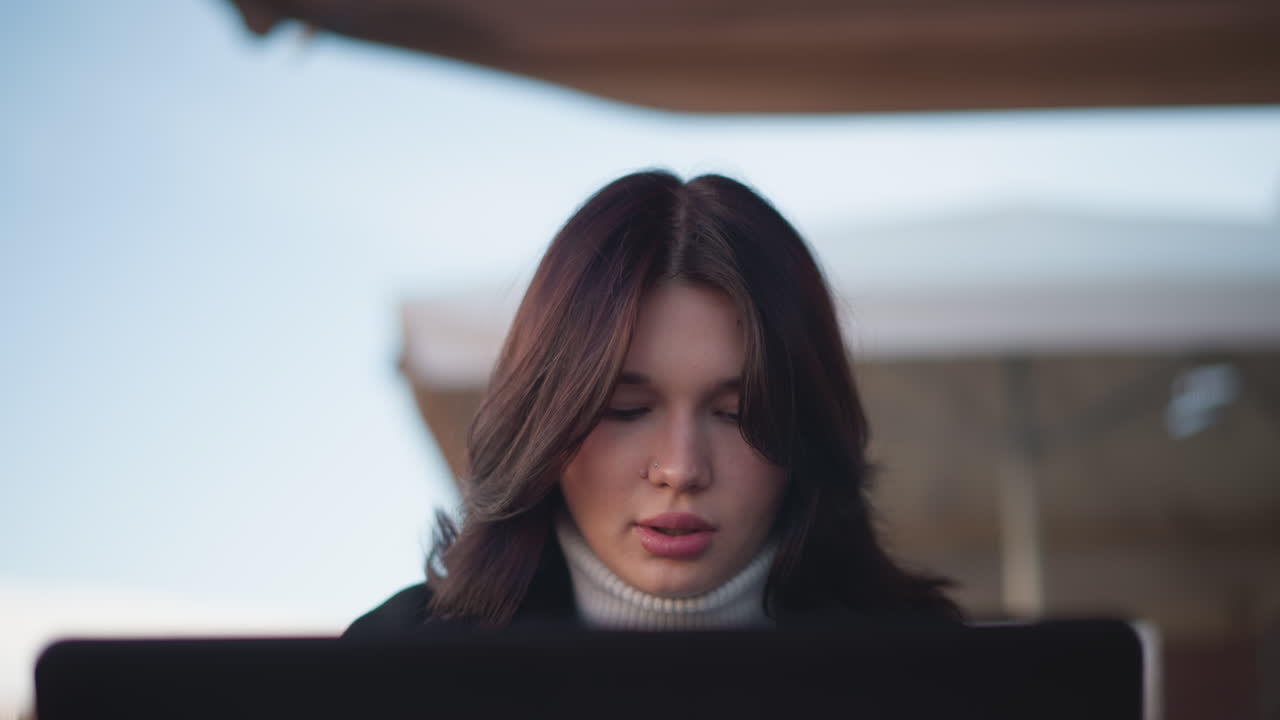 Lady seated with sober expression, wind gently swaying her soft brown hair as she faces something in front of her, soft lighting, blurred background
