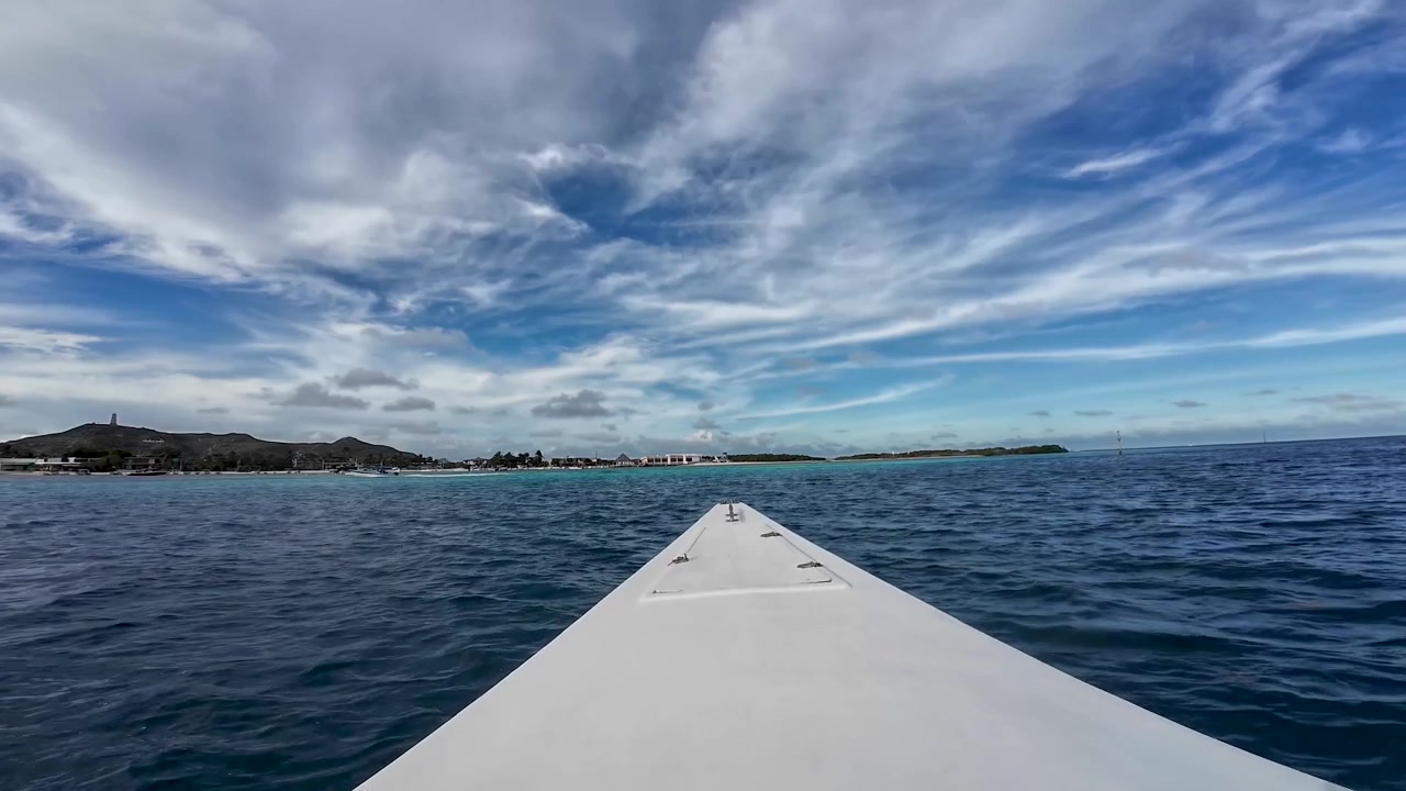 Navigazione POV attraverso le acque blu intenso dei Caraibi verso un'isola tropicale sotto un vasto cielo drammatico in un arcipelago remoto.