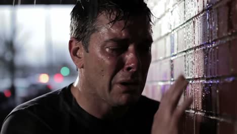 a man stands against a brick wall, rain pouring down as he expresses deep emotion. the setting is dark and moody, reflecting a sense of struggle and vulnerability in the late hours.