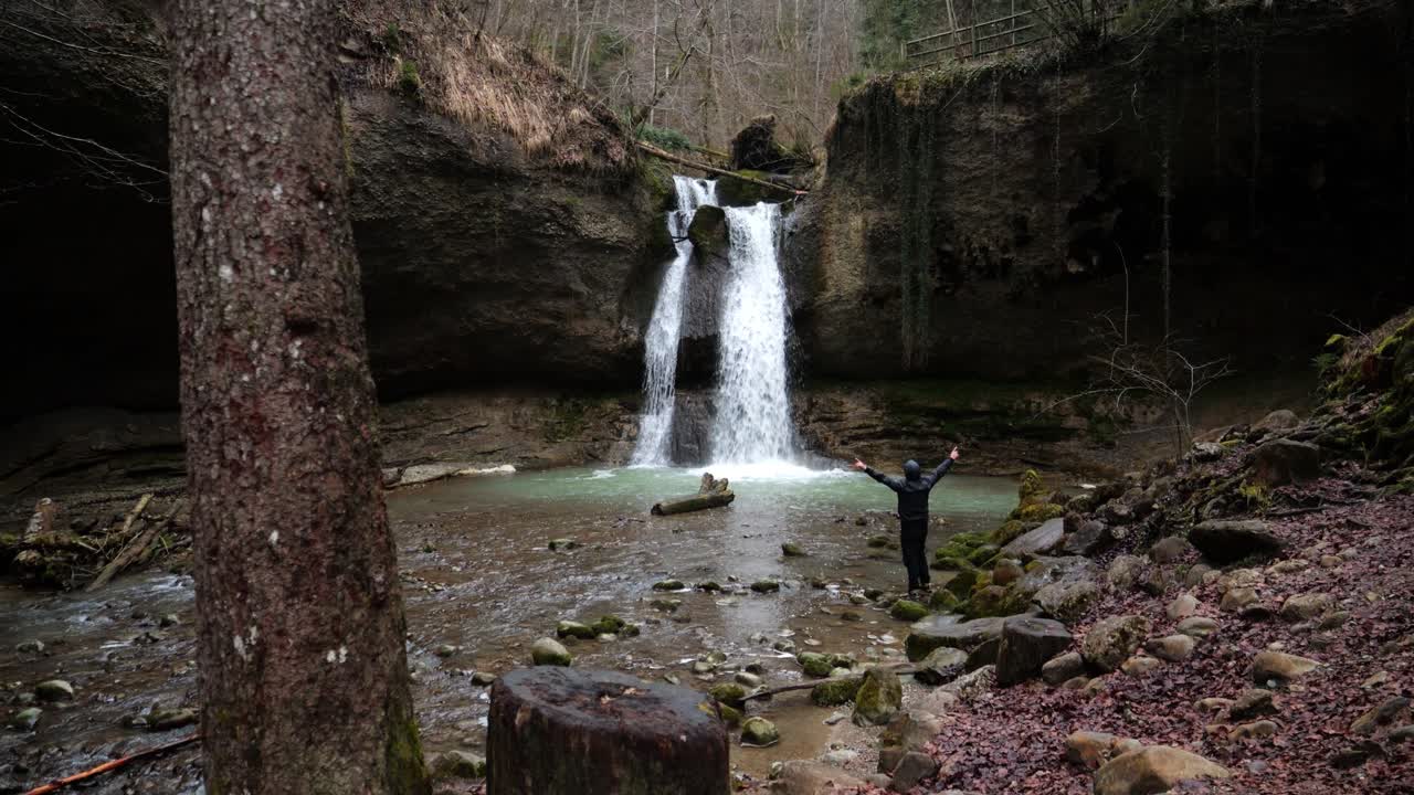 A man stands in a forest in front of a large waterfall, searching for hope. He stretches his arms up and contemplates the waterfall. Mystical natural landscape.