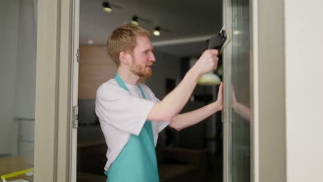 Side-view-of-a-confident-blond-male-cleaner-in-a-white-t-shirt-with-blue-aprons-cleaning-a-glass-wall-using-a-vacuum-cleaner-for-windows-in-a-modern-apartment