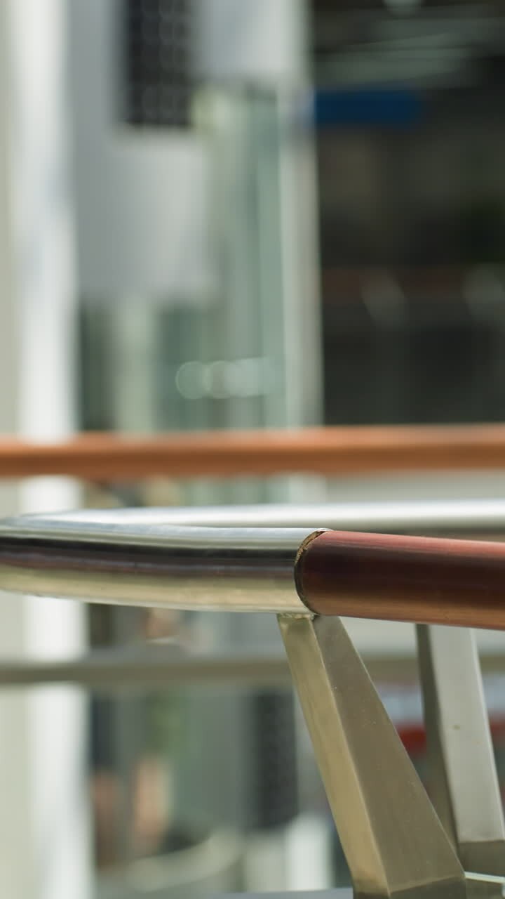 A dynamic mall scene captured from a first-person perspective with a handheld camera, featuring a blurred background. The focus is on sleek wooden handrails while a man and a woman pass by