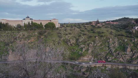 View-Of-The-Infantry-Academy-Building-Located-On-The-Opposite-Side-Of-River-Tagus-In-Toledo,-Spain-With-Vehicles-Driving-On-The-Road-Before-Sunset---high-angle-panning-shot