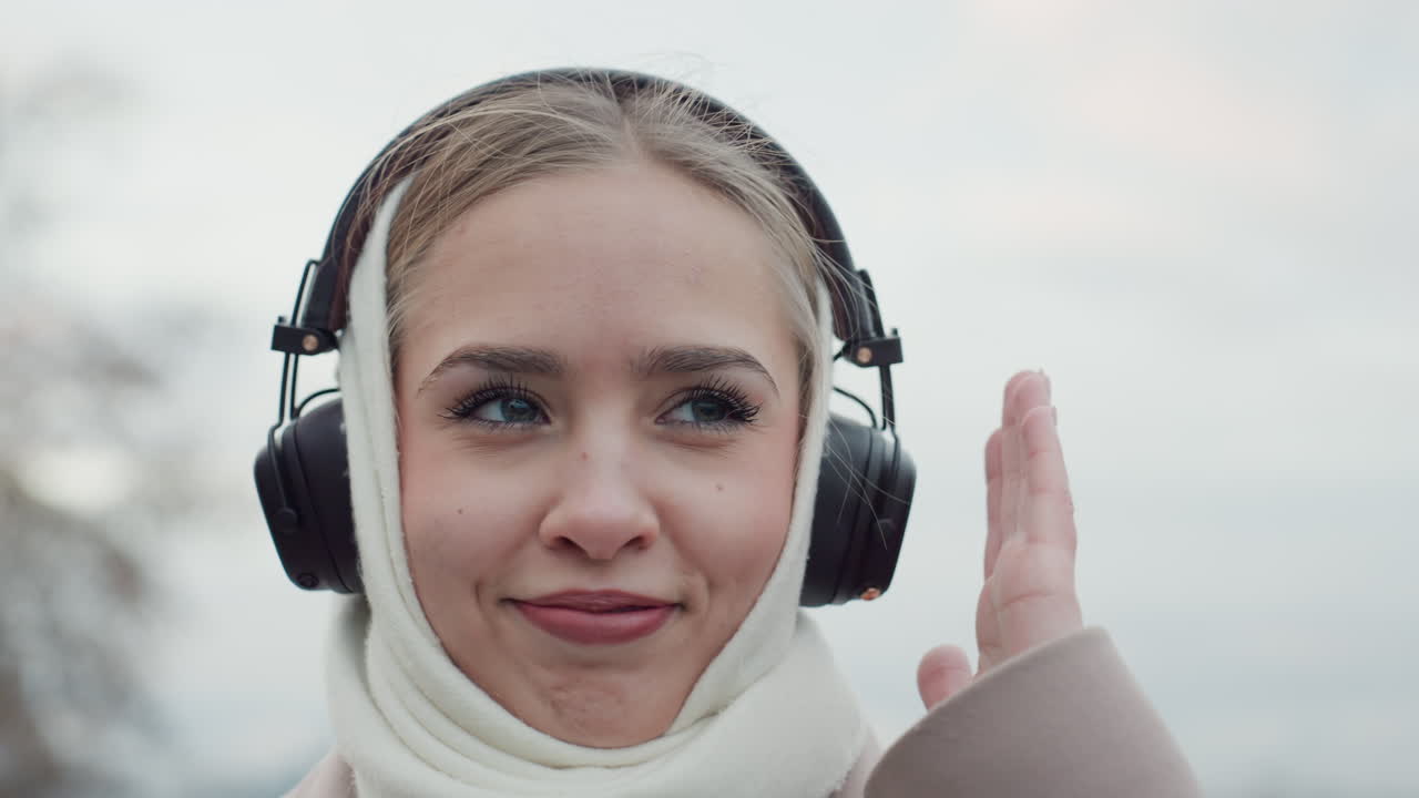 Overcast skyline with woman adjusting headphones. Woman in coat smiling and adjusting headphones outdoors. Cheerful woman wearing scarf adjusting her headphones against cloudy city background