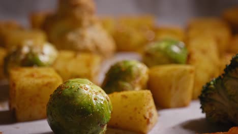 una toma macro que se inclina hacia abajo para revelar una col de bruselas cubierta con una capa de condimento en una bandeja cubierta con papel pergamino, una variedad de verduras y un trozo de pollo crudo rodean la col