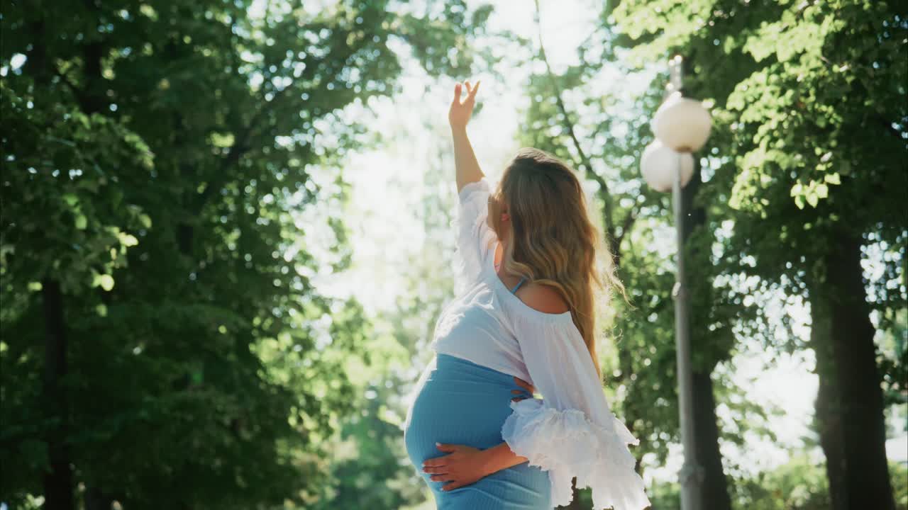 Mujer embarazada serena levantando el brazo hacia la luz del sol en el parque