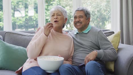 Happy-biracial-senior-couple-watching-tv-with-popcorn-and-having-fun-at-home