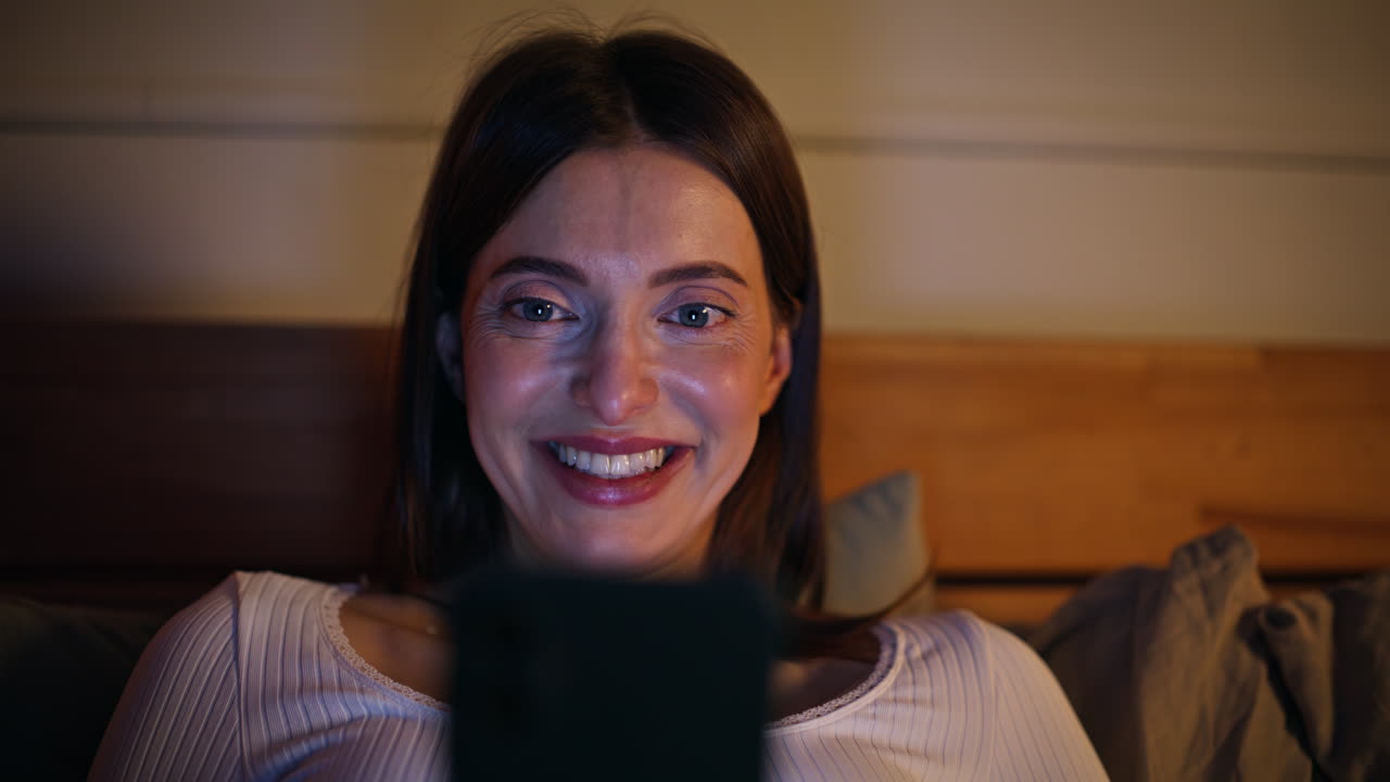 Donna sorpresa da un messaggio sul cellulare, sdraiata nel letto di casa, nel bagliore dello schermo del dispositivo, primo piano.