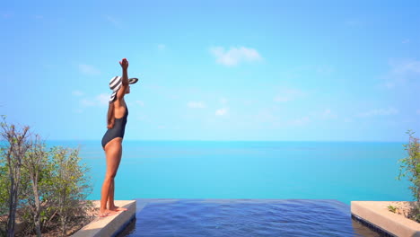 Mujer-En-Traje-De-Baño-Negro-Y-Sombrero-A-Rayas-Levanta-Los-Brazos-De-Pie-En-El-Borde-Pedregoso-De-La-Piscina-Infinita-En-La-Azotea-Disfrutando-Del-Impresionante-Paisaje-Marino