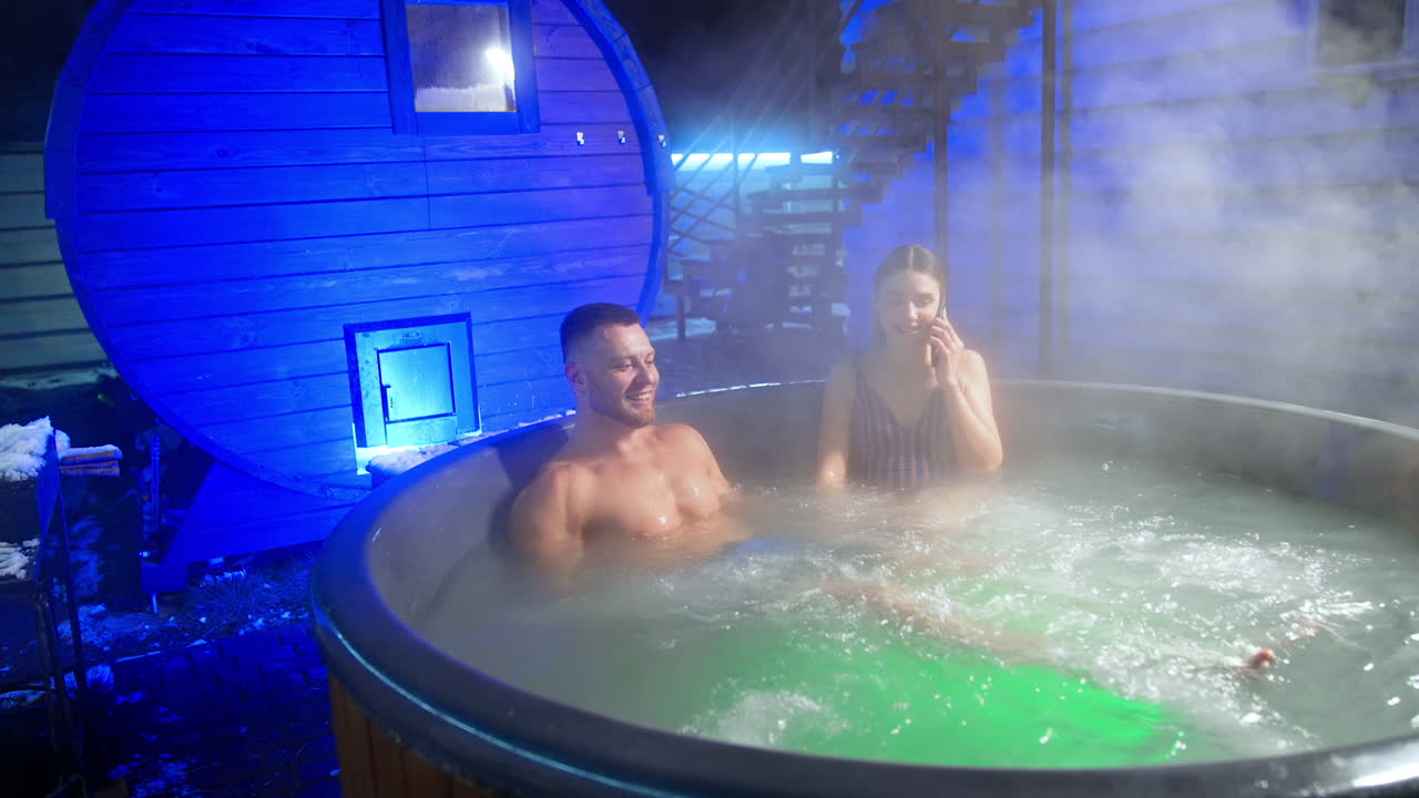 Smiling bearded man sits in hot tub moving his feet. Woman sitting beside talks on the phone.