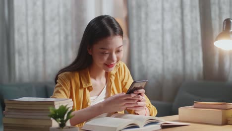 young asian female studying at home smiling and using smartphone while reading books on the table
