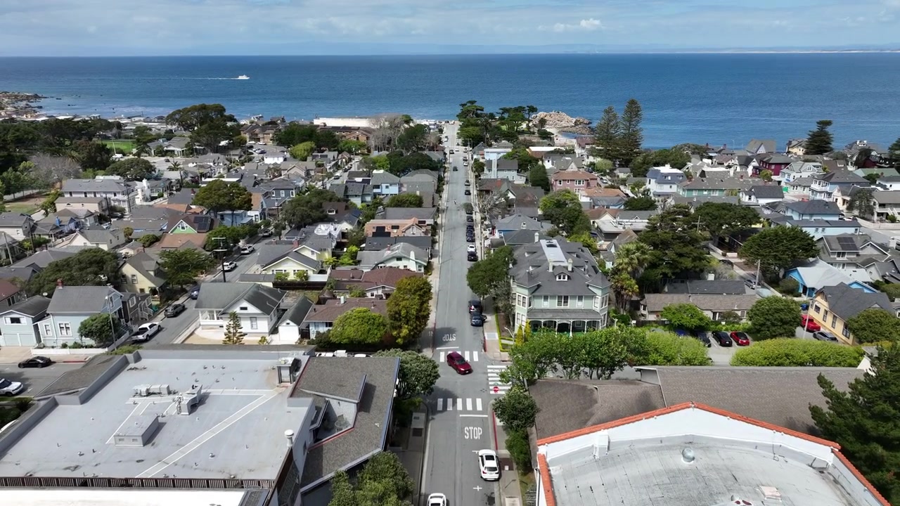 Filmatiske droneopptak fra luften som flyr bakover over et boligområde i Pacific Grove, California, og viser kystnære hjem, gater omkranset av trær, havutsikt og Lovers Point Park.