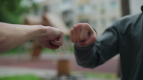 close-up of hands interacting in a playful knuckle bump, one person in a grey top extends their hand to connect with another, with a blurred background featuring residential buildings