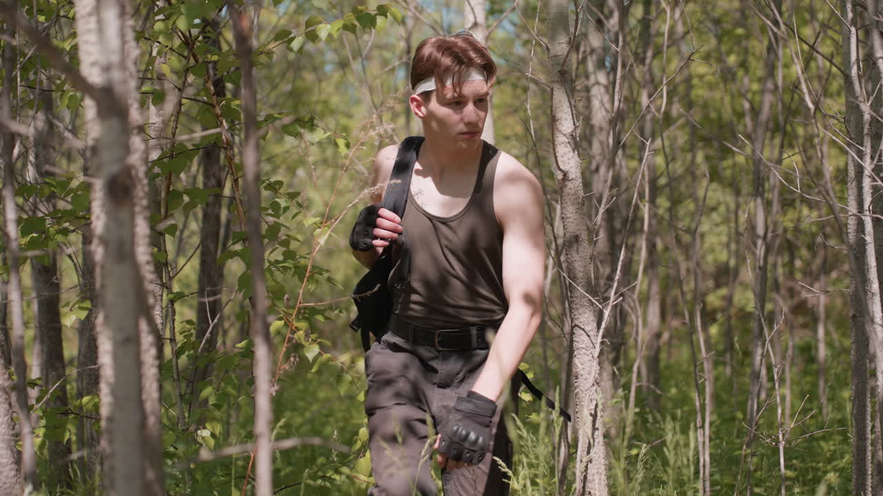 front view of tourist walking through dense green forest pushing aside bare branches while carrying black backpack and brushing past foliage under sunlight filtering through leafy canopy on trail