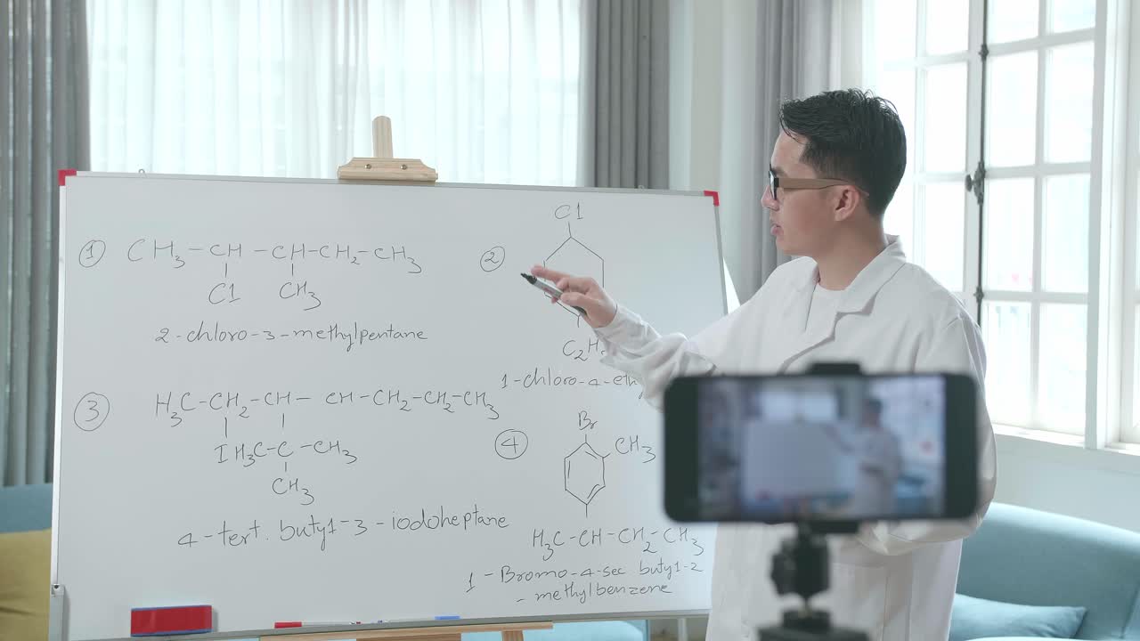 ครูชายชาวเอเชียสวมแว่นตากำลังถ่ายวิดีโอด้วยกล้องสมาร์ทโฟนขณะสอนวิชาเคมีที่บ้าน