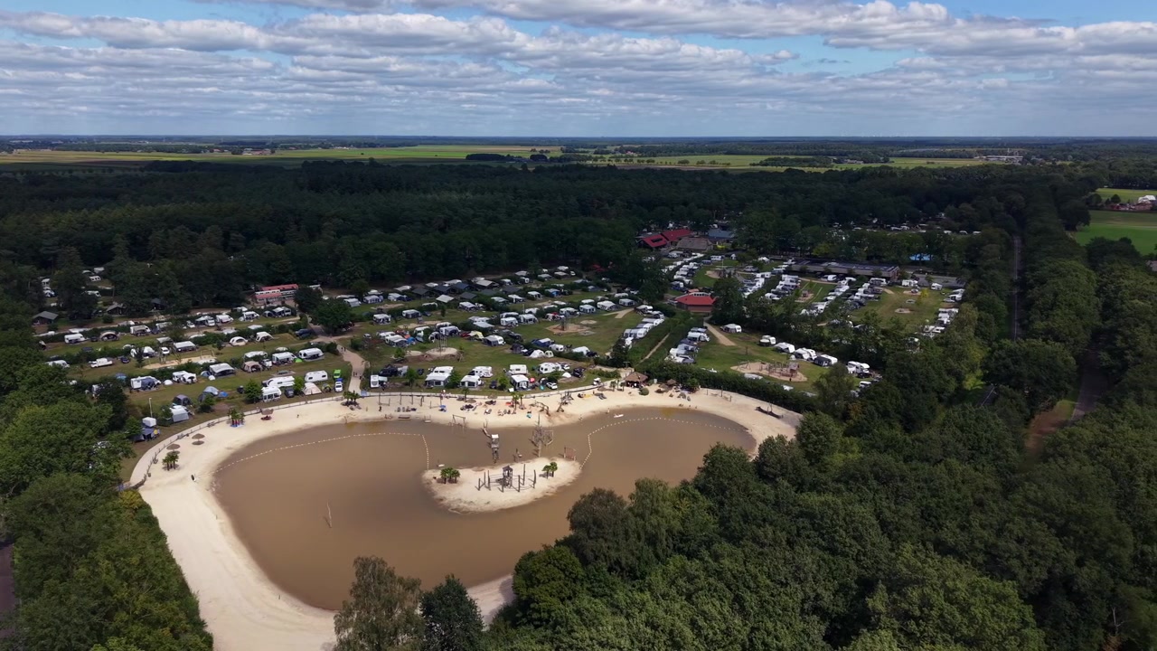 La spiaggia sabbiosa a forma di cuore di un lago circonda una piccola isola con altalene presso il campeggio De Kleine Wolf nei Paesi Bassi, file di camper costeggiano la radura nella foresta sotto un cielo nuvoloso. Ripresa aerea panoramica con drone