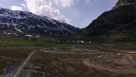 wide aerial drone shot flying over a vast, green alpine valley in montespluga, towards a small village nestled below majestic snow-capped mountains. italian alps, lombardy, italy