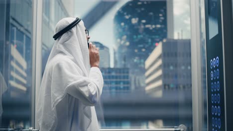 successful muslim businessman in traditional white kandura riding glass elevator to office in modern business center. man talking on a phone in a lift. saudi, emirati, arab businessman concept.