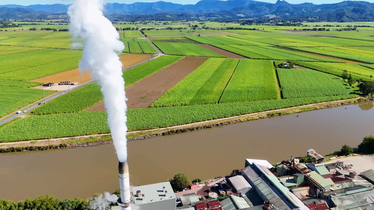 Drone footage captures a factory emitting steam amidst lush fields and a river under clear skies