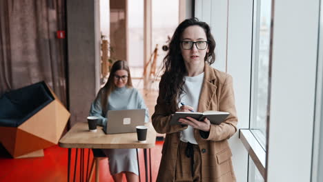 people working in the office. brunette lady stands at the window. business woman planning her day. blonde colleague working at laptop at backdrop.