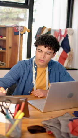 biracial male fashion designer sketches in studio with laptop, vertical video, slow motion