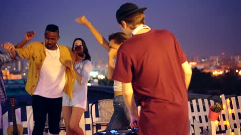 cheerful male deejay is working with mixing console and dancing while group of friends caucasian and african american are having fun at rooftop party.