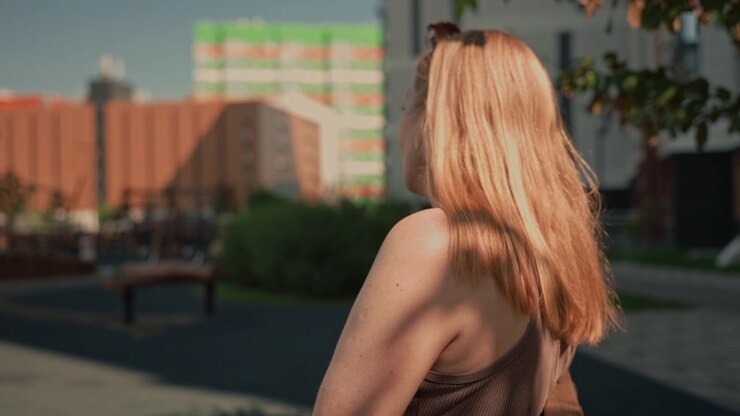 Una donna con lunghi capelli biondi sta all'aperto in un parco pubblico, di spalle alla macchina fotografica e guarda in lontananza. La donna indossa un top marrone senza maniche mentre la luce solare proietta ombre sul suo braccio.