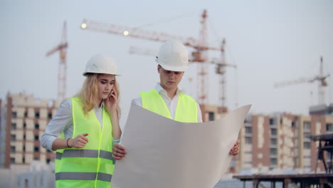 Dos-Colegas-Ingenieros,-Hombre-Y-Mujer,-Discutiendo-El-Dibujo-En-El-Fondo-De-Edificios-Y-Grúas,-Una-Mujer-Hablando-Por-Teléfono-Con-El-Jefe