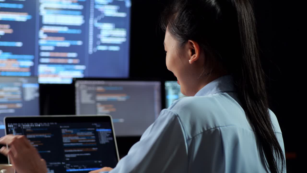 vista de atrás de una programadora asiática bebiendo café mientras escribe código en una computadora portátil usando múltiples monitores que muestran la base de datos en los escritorios de ventana de terminal en la oficina