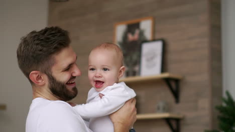 Loving-father-in-white-t-shirt-hugs-the-baby-and-kisses-the-baby-laughs-and-smiles-on-the-background-of-the-Christmas-tree-and-garlands